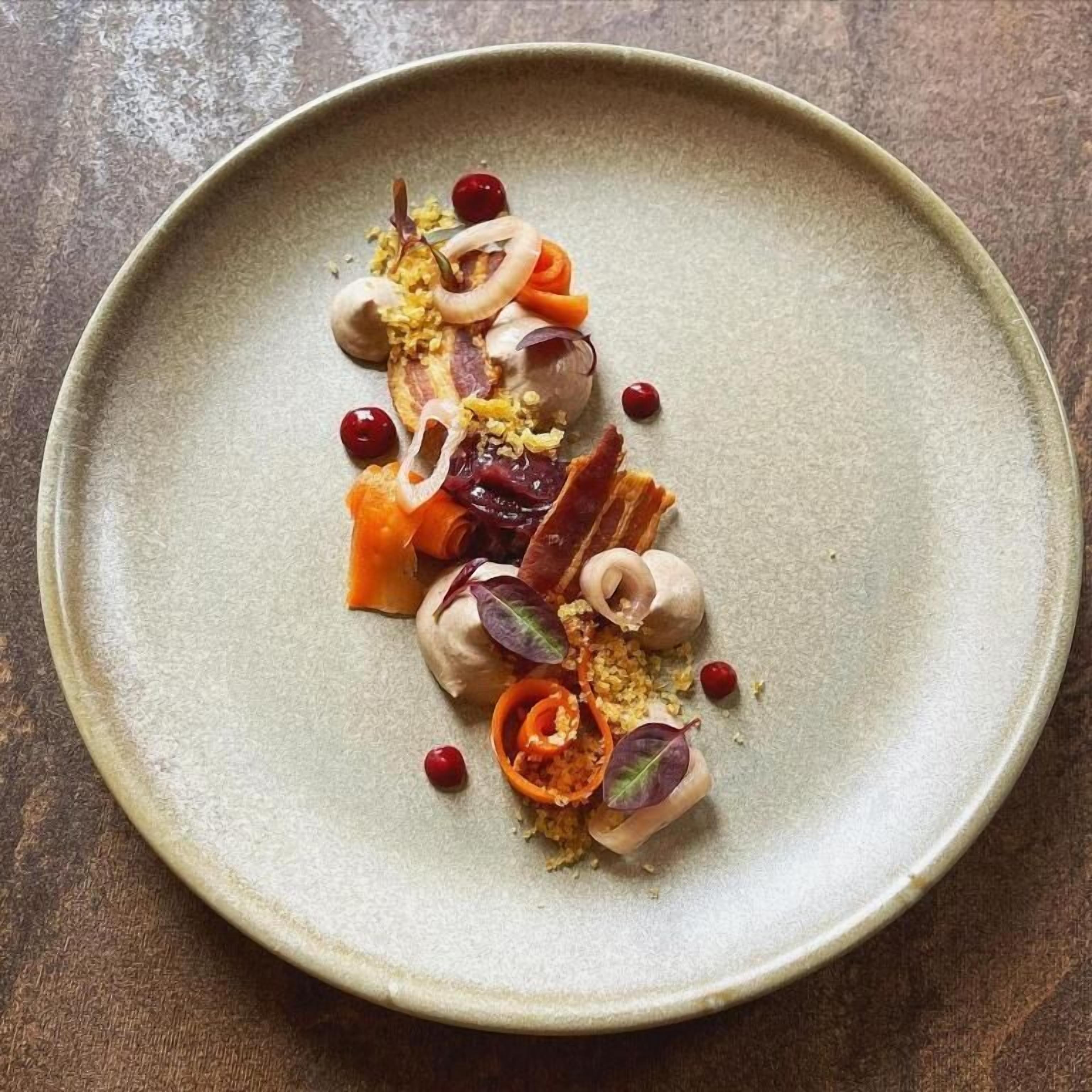 A round ceramic plate with a minimalist design, featuring a variety of food items including what appears to be a type of meat, vegetables, and possibly some nuts or seeds.