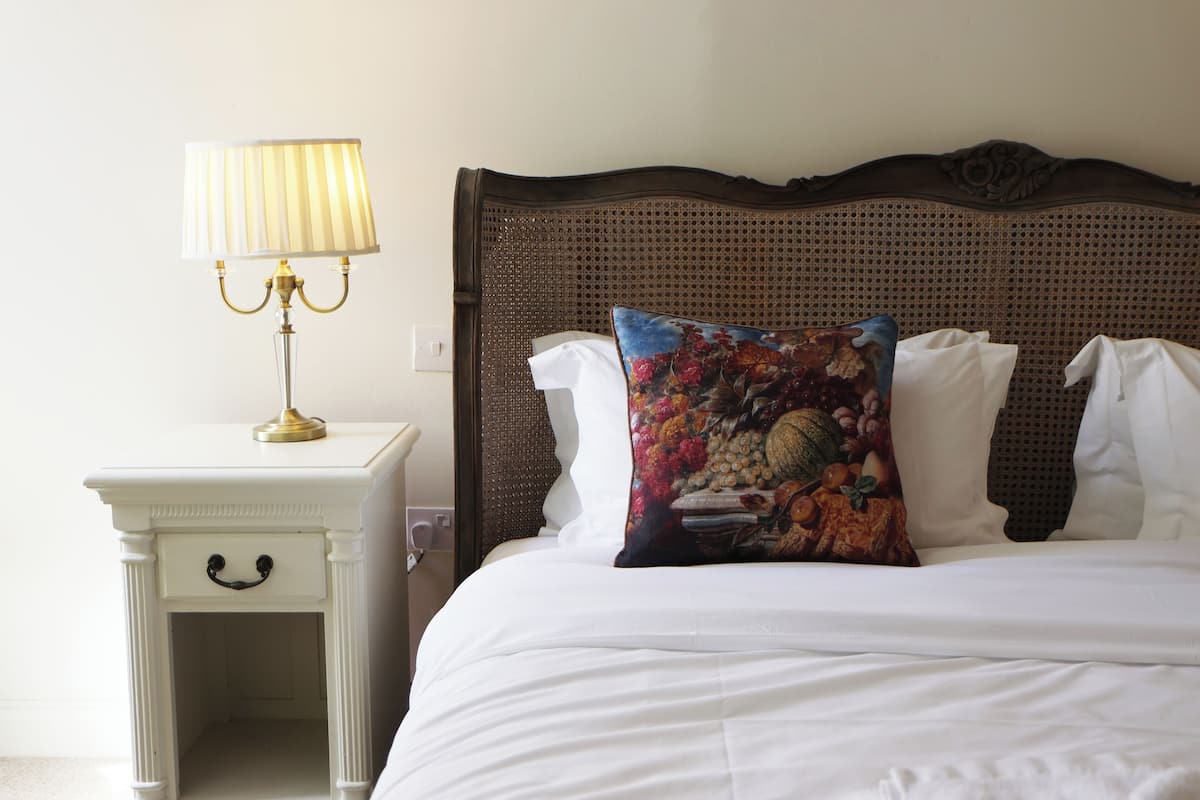 A neatly made bed with white linens and a floral pillow, a white bedside table with a lamp, and a dark wooden headboard.