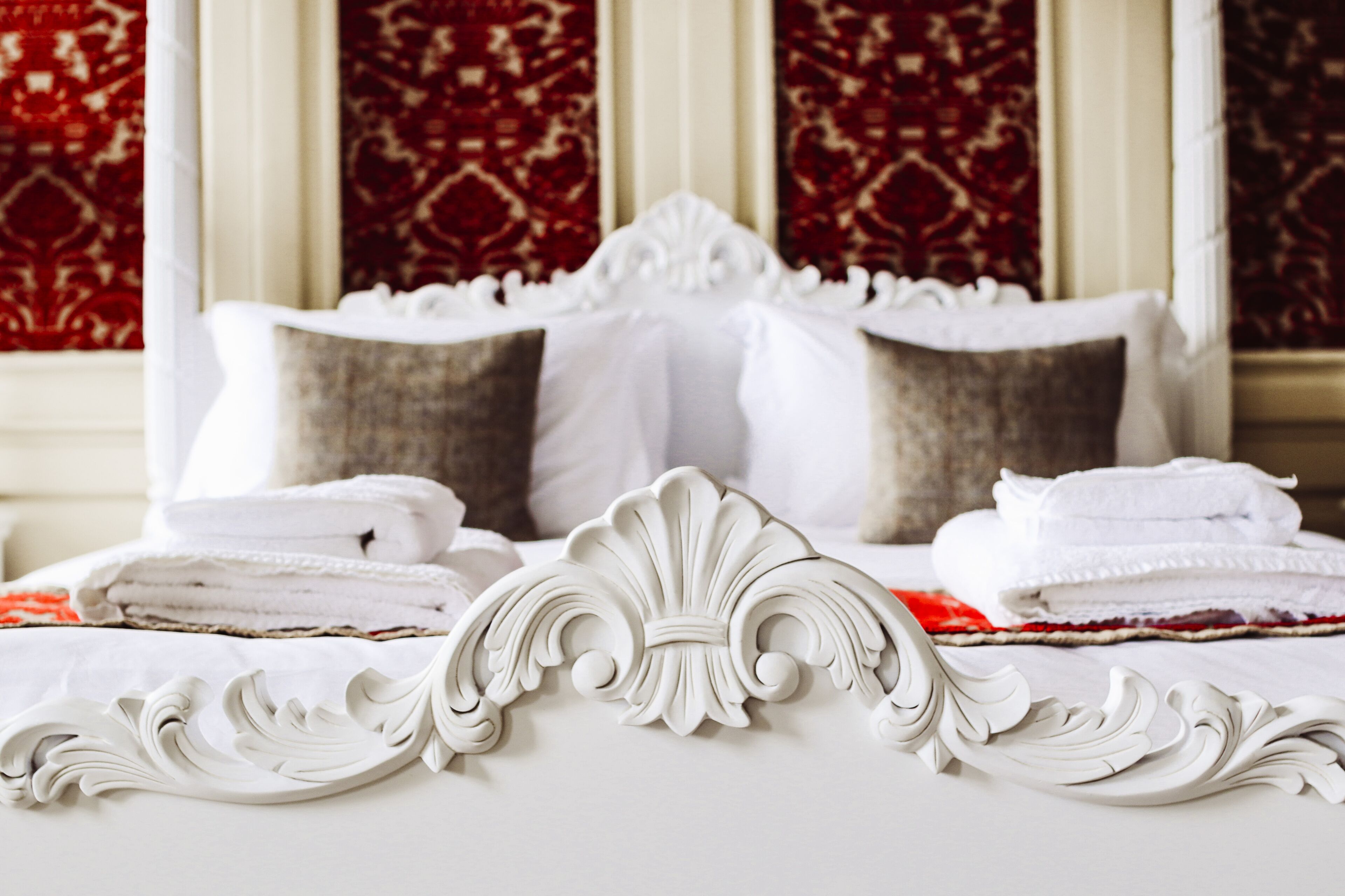 A neatly made bed with white linens and decorative headboard, set against a backdrop of patterned red curtains.