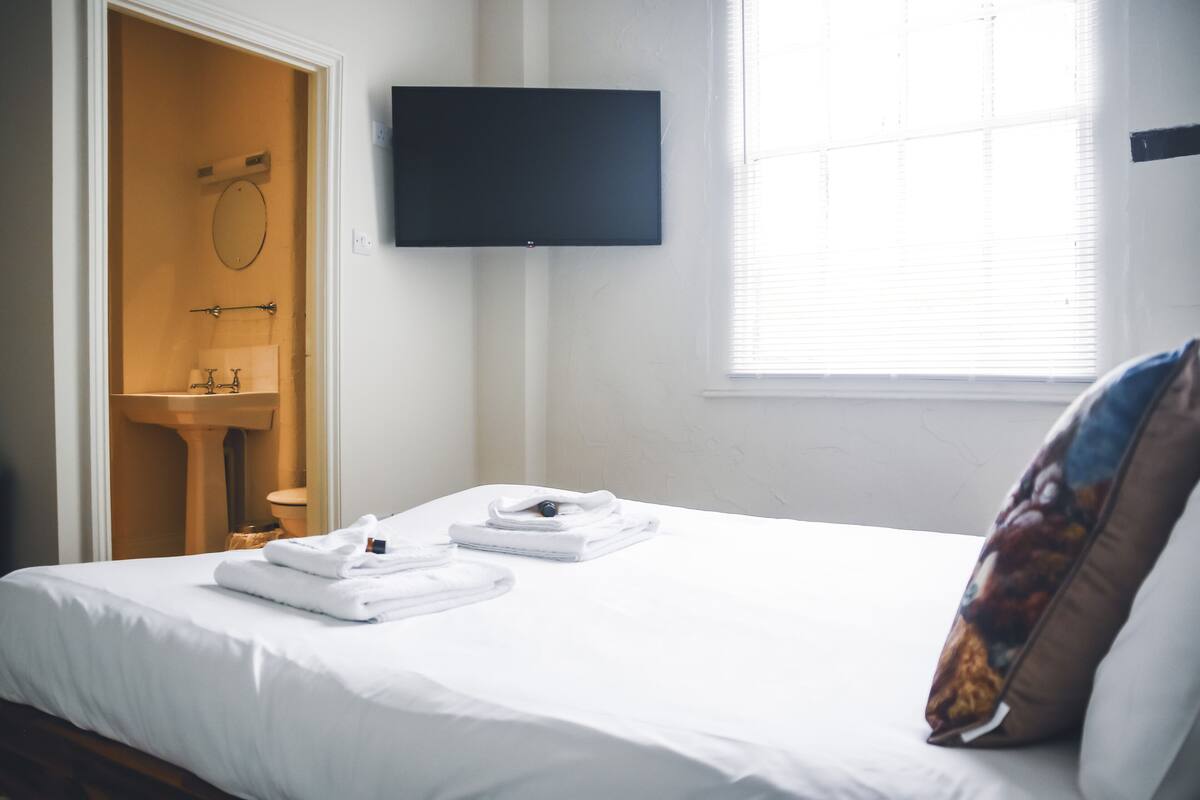 A hotel room with a bed, a television mounted on the wall, and a window with blinds.