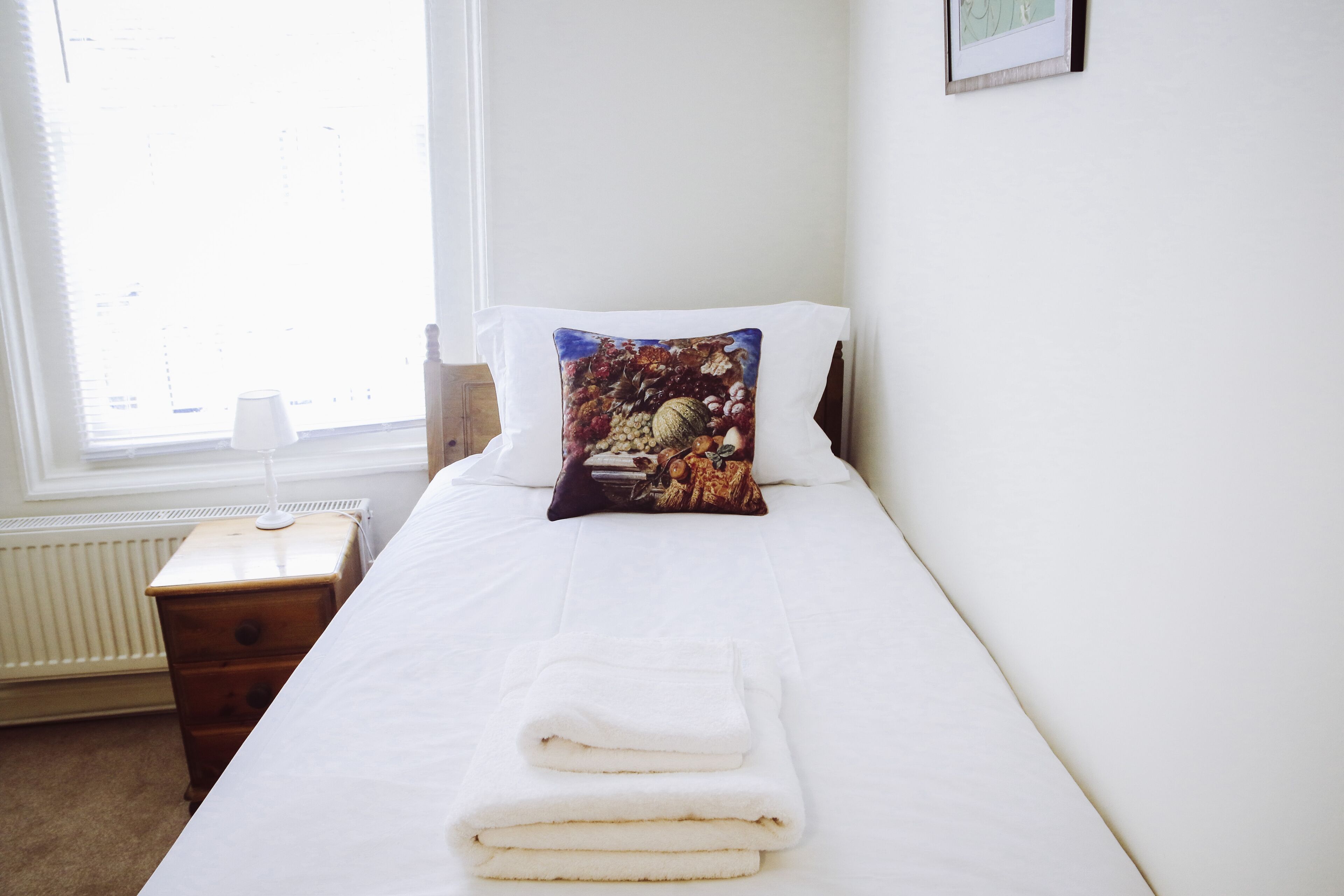 A neatly made bed with a white duvet and a decorative pillow, a wooden nightstand with a lamp, and a window with blinds.