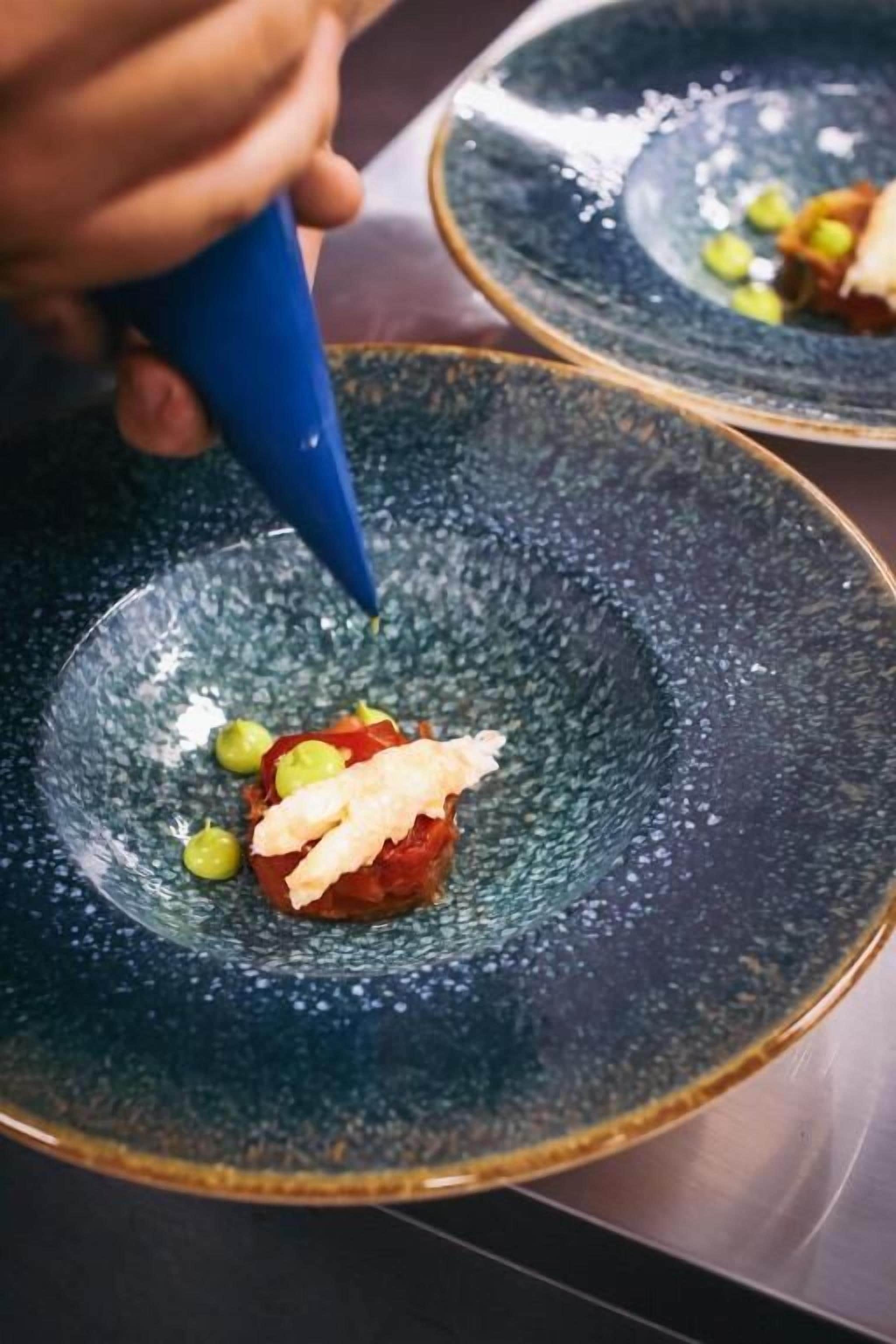 Un xef utilitzant una bossa de pastisseria blava per afegir salsa a un plat de ceràmica blau fosc.
