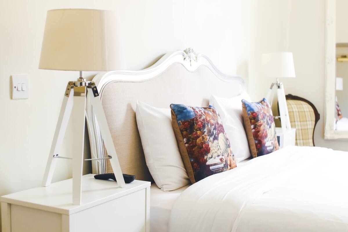A neatly made bed with white bedding and decorative pillows, a bedside table with a lamp, and a wall socket.