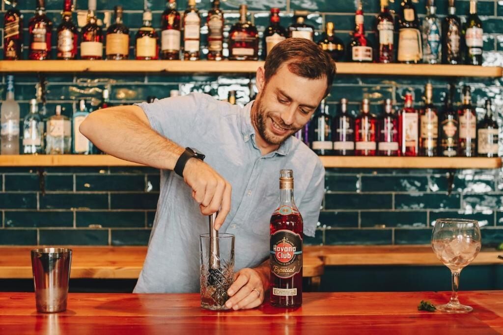 En bartender laver en drink ved en bar med hylder fyldt med flasker spiritus i baggrunden.
