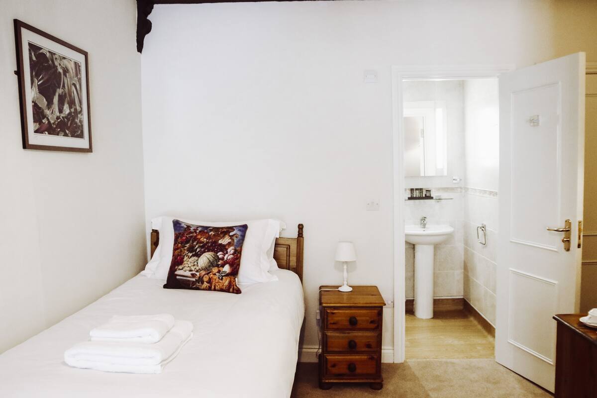 A bedroom with a bed, a nightstand, a bedside lamp, a bathroom with a sink, and a door leading to another room.