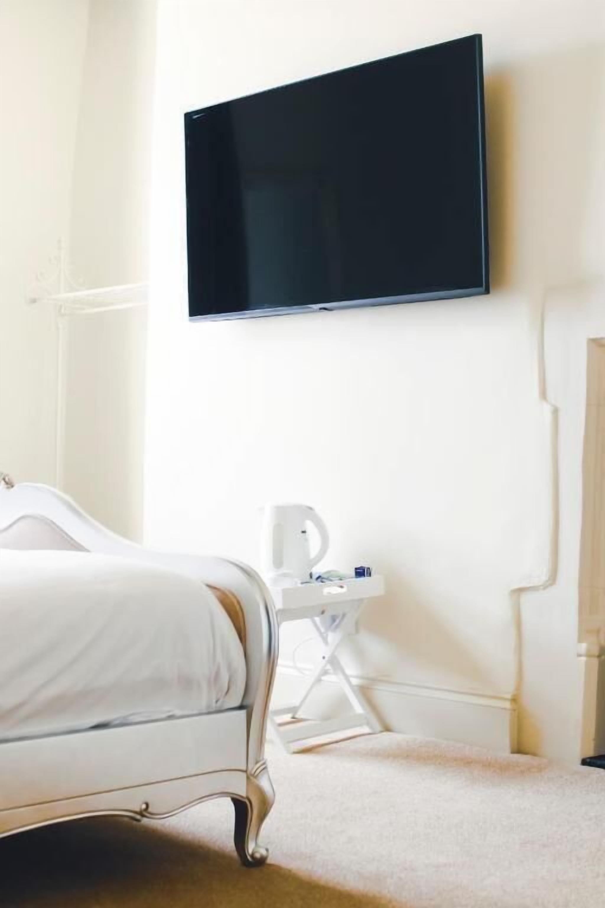 Una habitación de hotel con una cama, un televisor de pantalla plana en la pared, una mesita de noche con lámpara y una silla blanca.