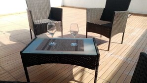 Een klein terras met rieten stoelen en een tafel met glazen blad, met twee wijnglazen.