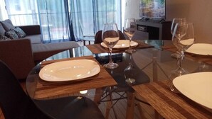 Een eethoek met een glazen tafel, witte borden en wijnglazen. Achtergrond is een bank en een televisie.