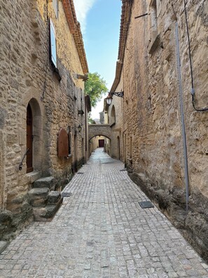 Une ruelle étroite pavée, bordée de bâtiments historiques en pierre, dotés d’entrées en arc de cercle et d’une arche centrale menant à une cour intérieure.