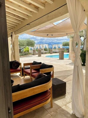 A covered outdoor seating area with wooden furniture and a view of a pool and landscape.