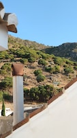 A building with a white exterior and a brown roof, overlooking a hillside with sparse trees and rocky terrain.