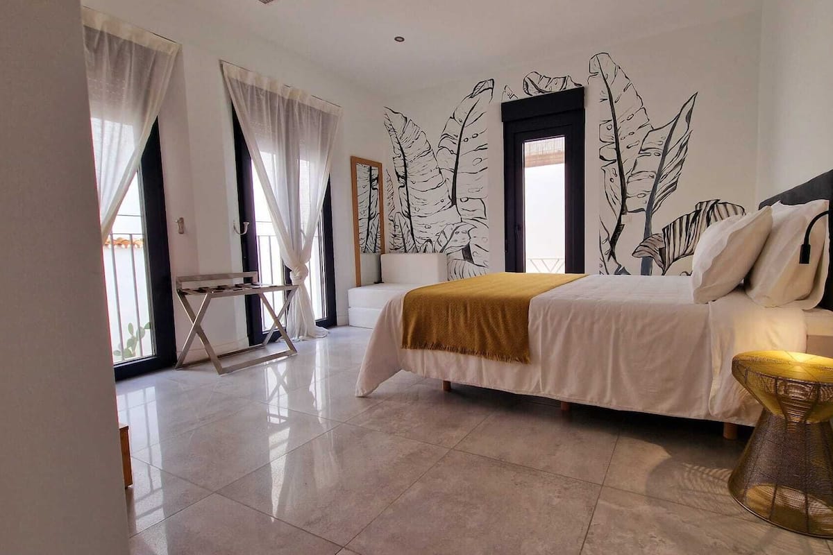 A modern bedroom with a bed, a small table, and a chair. There are large windows with sheer curtains, and a wall mural of leaves.