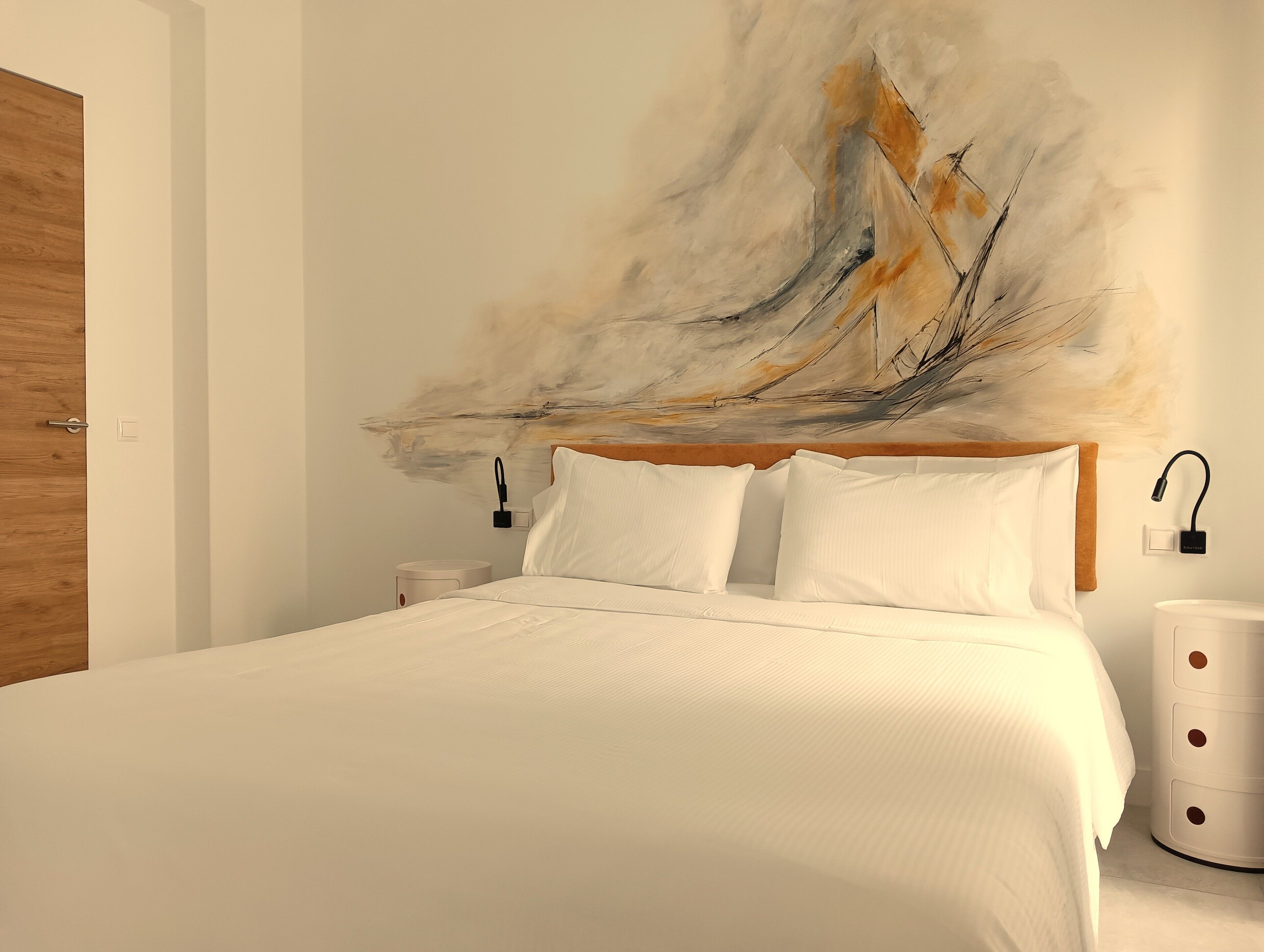 Una cama con sábanas blancas impecables en una habitación moderna con cabecera de madera, una lámpara de pared y una mesita de noche blanca.