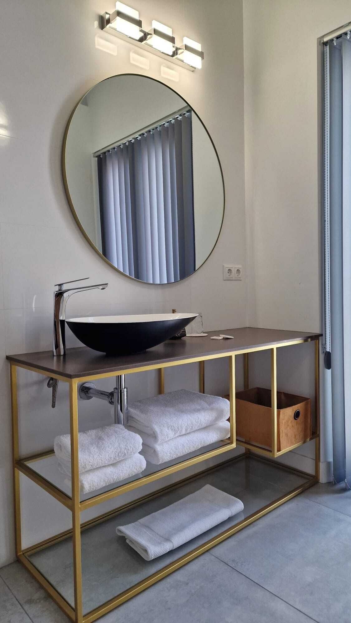 Een moderne badkamer met een ronde spiegel, een wastafel op voet en een gouden wastafelmeubel met planken voor handdoeken.