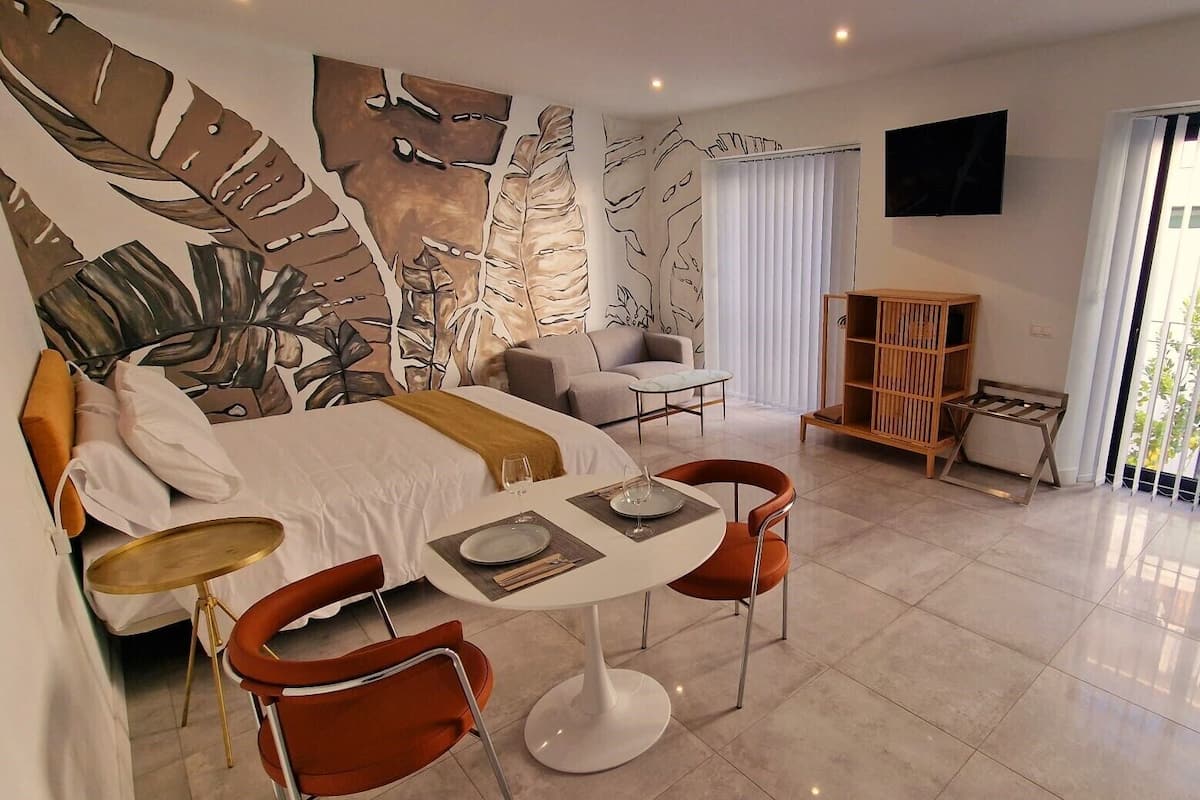 A modern hotel room with a large bed, a sitting area, a dining table with chairs, and a TV mounted on the wall.