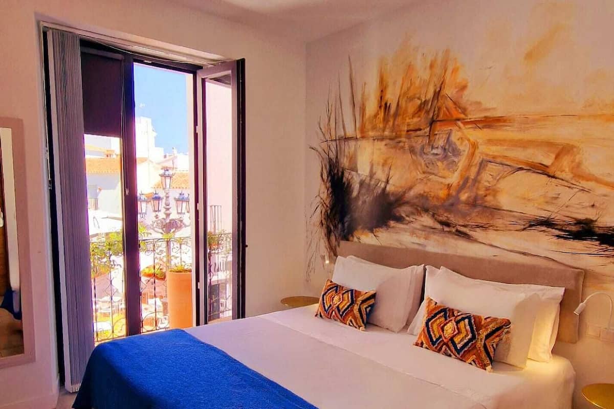 A hotel room with a large bed, a painting on the wall, and a balcony with a view of the city.