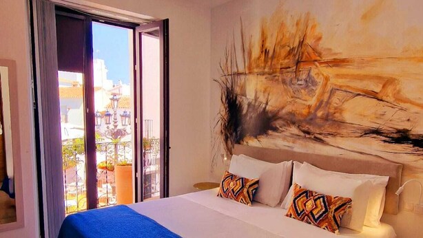 A hotel room with a large bed, a painting on the wall, and a balcony with a view of the city.