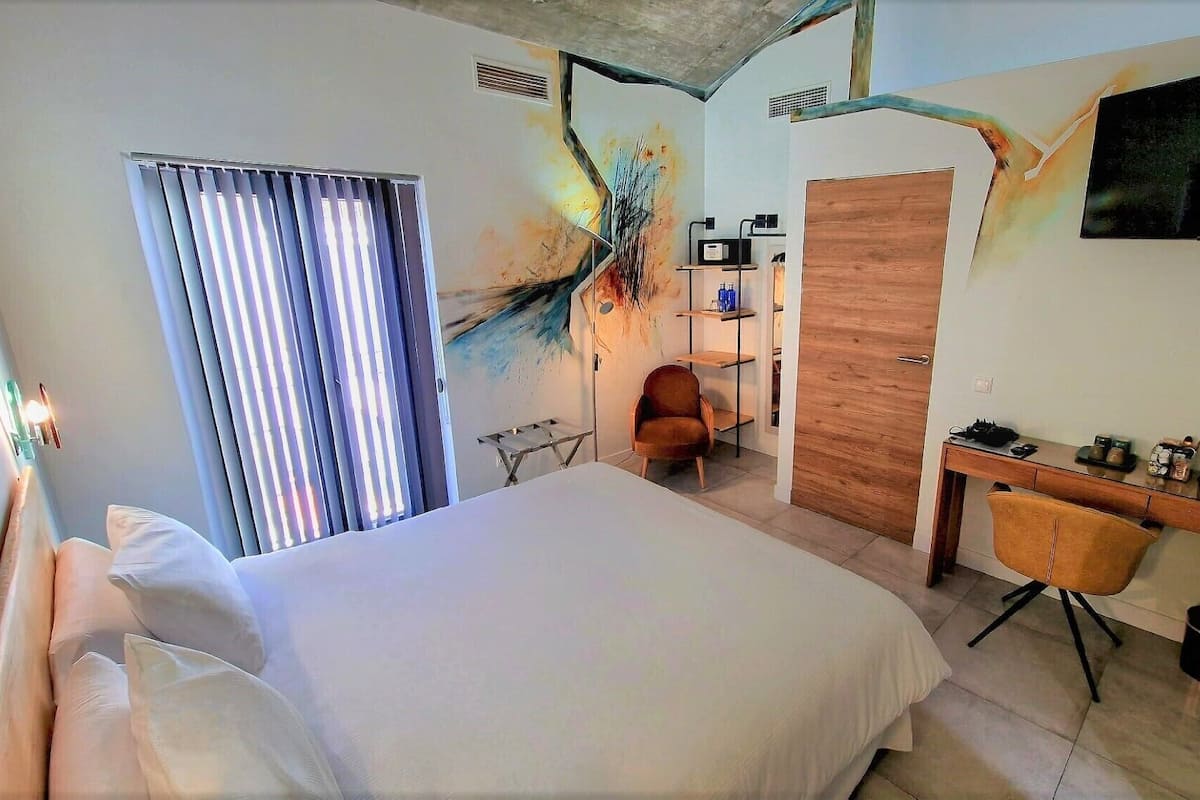 A hotel room with a large bed, a desk with a chair, a TV mounted on the wall, and a window with blinds.