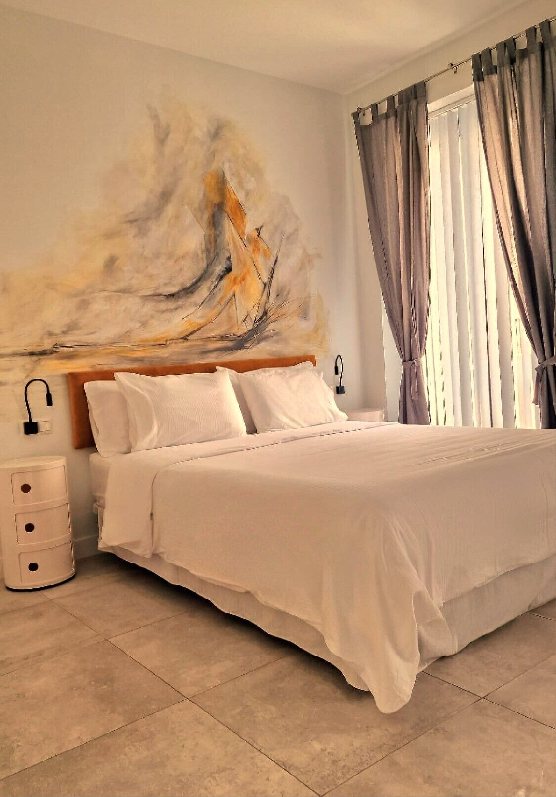 A bedroom with a large bed, a bedside table, a wall-mounted lamp, and a window with curtains.