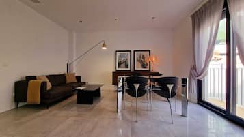 A modern living room with a sofa, coffee table, and dining area with chairs. There are two framed pictures on the wall, a floor lamp, and a large window with curtains.