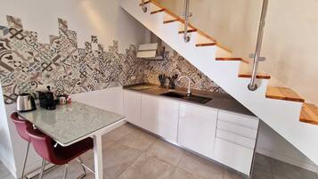 Eine moderne Küche mit weißen Schränken, einem Esstisch mit Glasplatte und einem roten Stuhl. Eine Holztreppe führt zu einer Erkerzone.