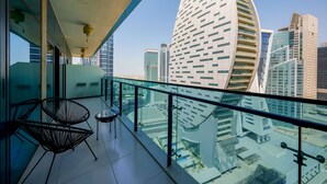 A modern hotel balcony with glass railings, two black wireframe chairs, and a view of a cityscape with distinctive architecture. - Tanin - Apartment Amidst Lively Area With Pool and Balcony (Dubai)