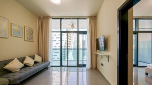 A modern living room with a grey sofa, a flat-screen TV on a black stand, and a large window with curtains. - Tanin - Apartment Amidst Lively Area With Pool and Balcony (Dubai)