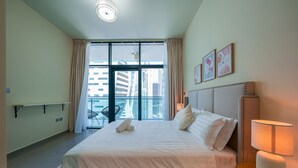 A hotel room with a large bed, a bedside lamp, a window with curtains, and a view of buildings. - Tanin - Apartment Amidst Lively Area With Pool and Balcony (Dubai)