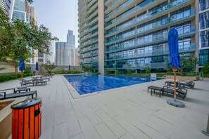 Un área de piscina moderna al aire libre con sillones de descanso y sombrillas, rodeada de edificios altos.