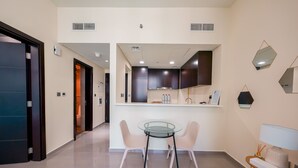 A modern kitchen with dark cabinets, a glass-top table, and two chairs. There is a small island with a sink and a mirror above it. - Tanin - Apartment Amidst Lively Area With Pool and Balcony (Dubai)