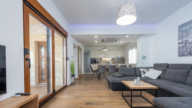 A modern living room with a grey sofa, a wooden coffee table, and a TV on a wooden stand. There is a sliding glass door leading to a kitchen area. - Villa Roza Zadarvillas (Policnik)