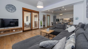 A modern living room with a large gray sofa, a wooden coffee table, and a TV on a wooden shelf. There is a kitchen in the background with a dining area visible through an open door. - Villa Roza Zadarvillas (Policnik)