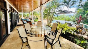 Une terrasse extérieure avec une table ronde en verre et des chaises, entourée de plantes tropicales et offrant une vue sur une piscine.