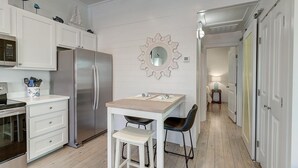A modern kitchen with white cabinetry, a stainless steel refrigerator, a dining table with two chairs, and a decorative wall mirror. - Pier 15: 2 Pools, Outdoor Dining, Covered Parking (Port Aransas)