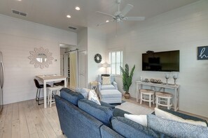 A spacious living room with a large blue sofa, a dining area with a white table, and a TV mounted on the wall. - Pier 15: 2 Pools, Outdoor Dining, Covered Parking (Port Aransas)