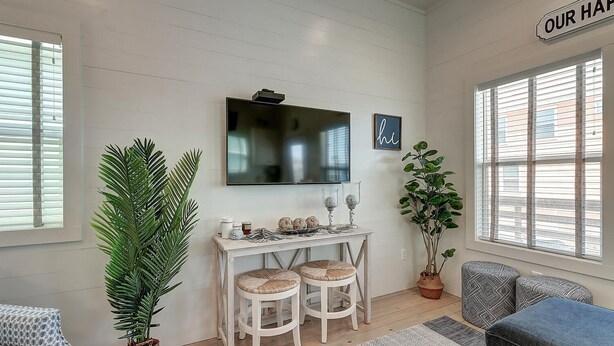 A cozy living room with a white wall, a flat-screen TV, a small table with two chairs, a large potted plant, and a blue sofa. - Pier 15: 2 Pools, Outdoor Dining, Covered Parking (Port Aransas)