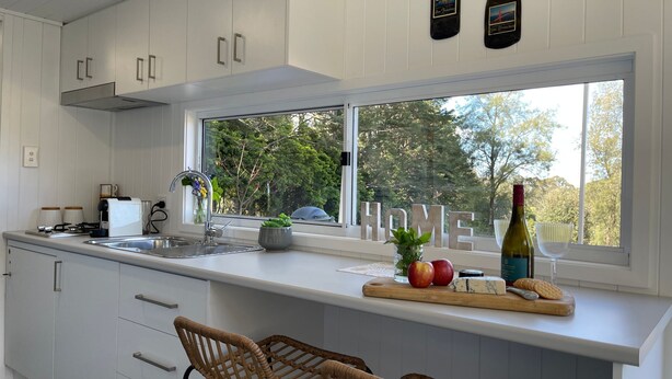 A modern kitchen with white cabinetry, a large window with a view of trees, a wooden cutting board with fruits and cheese, and a bottle of wine.