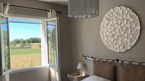 A bedroom with a bed, a bedside table with a lamp, a circular wall decoration, and a view of a field through a window. - Villa-mazet (Argilliers)