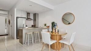 A modern kitchen with white cabinets, a built-in oven, and a stainless steel refrigerator. There is a dining area with a round wooden table and white chairs. - Beachside Haven Modern Townhouse with Pool (Casuarina)