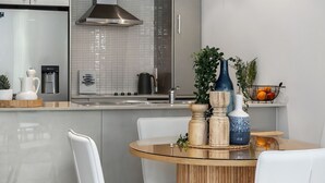 A modern kitchen with a stainless steel refrigerator, a water dispenser, and a kettle. There is a dining table with a glass top and wooden legs, surrounded by white chairs. - Beachside Haven Modern Townhouse with Pool (Casuarina)