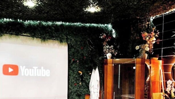 Ett spaområde med en stor skärm som visar YouTubes logotyp, en svart lädersoffa, en träbänk, en bubbelpool och ett träskåp med en vas med blommor.