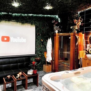 Un área de spa con una pantalla grande mostrando el logotipo de YouTube, un sofá de cuero negro, un banco de madera, una bañera de hidromasaje y un mueble de madera con un jarrón de flores.