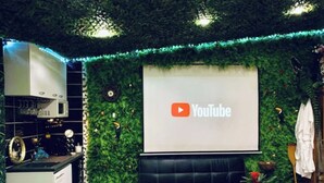 Ett modernt vardagsrum med en YouTube-logotyp på väggen, en svart soffa, ett litet bord med dekorativa föremål och en köksdel med en vit köksapparat.