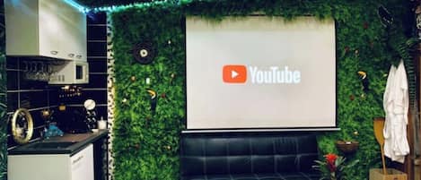 A modern living room with a YouTube logo on the wall, a black sofa, a small table with decorative items, and a kitchen area with a white appliance.