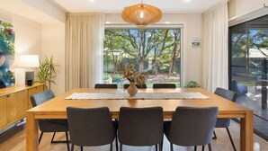 A modern dining room with a wooden table, grey chairs, a hanging light fixture, and a view of trees through large windows. - Luxurious Privacy in Exquisite Home, Walk to Golf (Cowes)