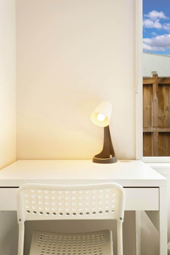 A white desk with a lamp, a white chair, and a window with a view of a wooden fence.