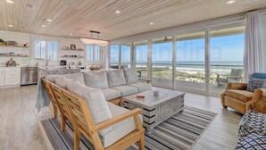A spacious living area with a large sofa, wooden chairs, and a coffee table. It features a kitchen with white cabinets and a view of the beach.