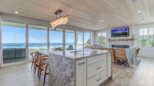 A modern kitchen with a large island, wooden ceiling, and large windows overlooking the ocean.