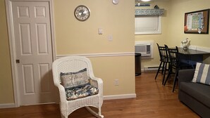 Een woonkamer met een rieten stoel, een eettafel met stoelen, een grijze bank en een wandklok.