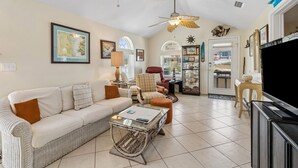 A living room with a sofa, armchairs, a coffee table, a TV, ceiling fans, and a door leading to another room. - 534 N. Fletcher Ave. #1 2 Bedroom Cottage by RedAwning (Fernandina Beach)
